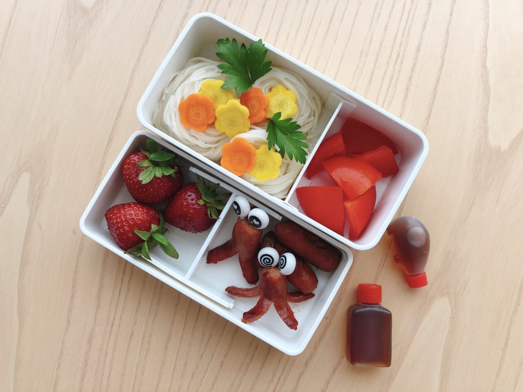 Bento box lunch features hot dogs cut into octopuses, strawberries, tomato wedges, and somen noodles.