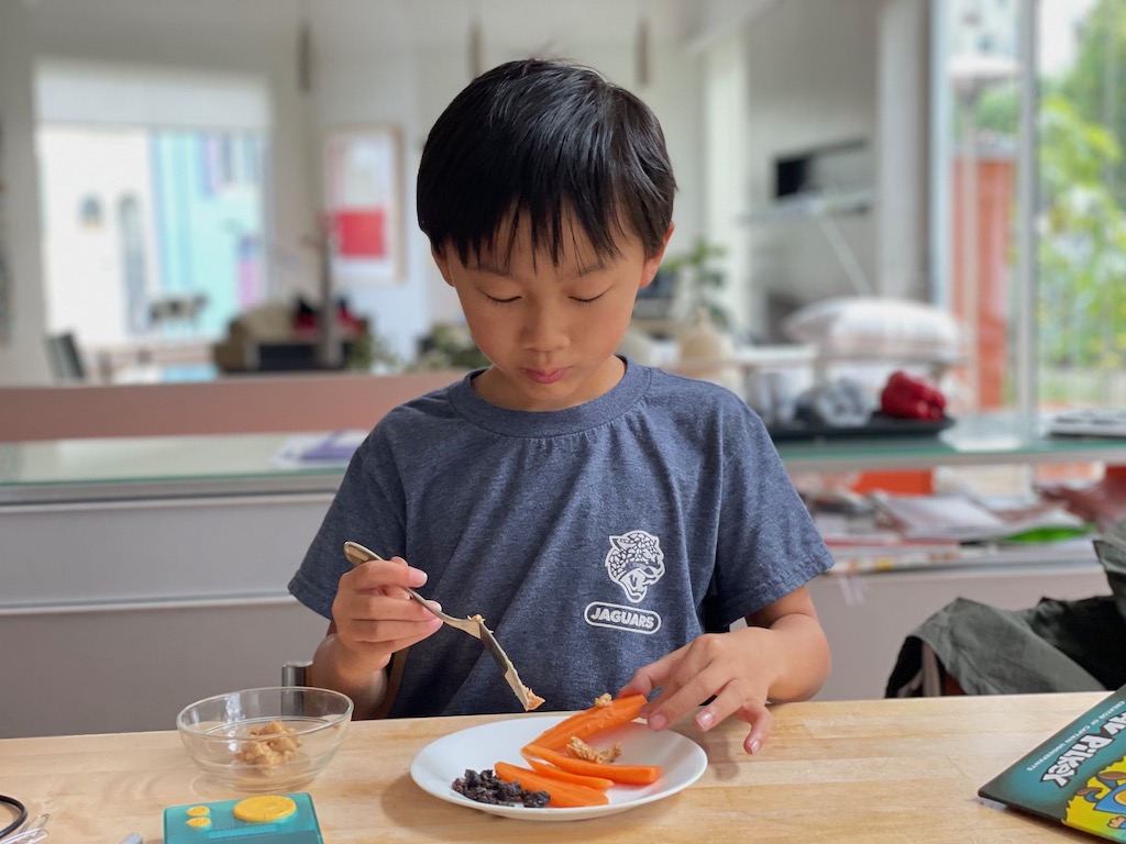 Child spreads peanut butter on carrot sticks for ants on a log.