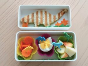 A brontosaurus bento box lunch is made by cutting a grilled cheese sandwich with a dinosaur cookie cutter.