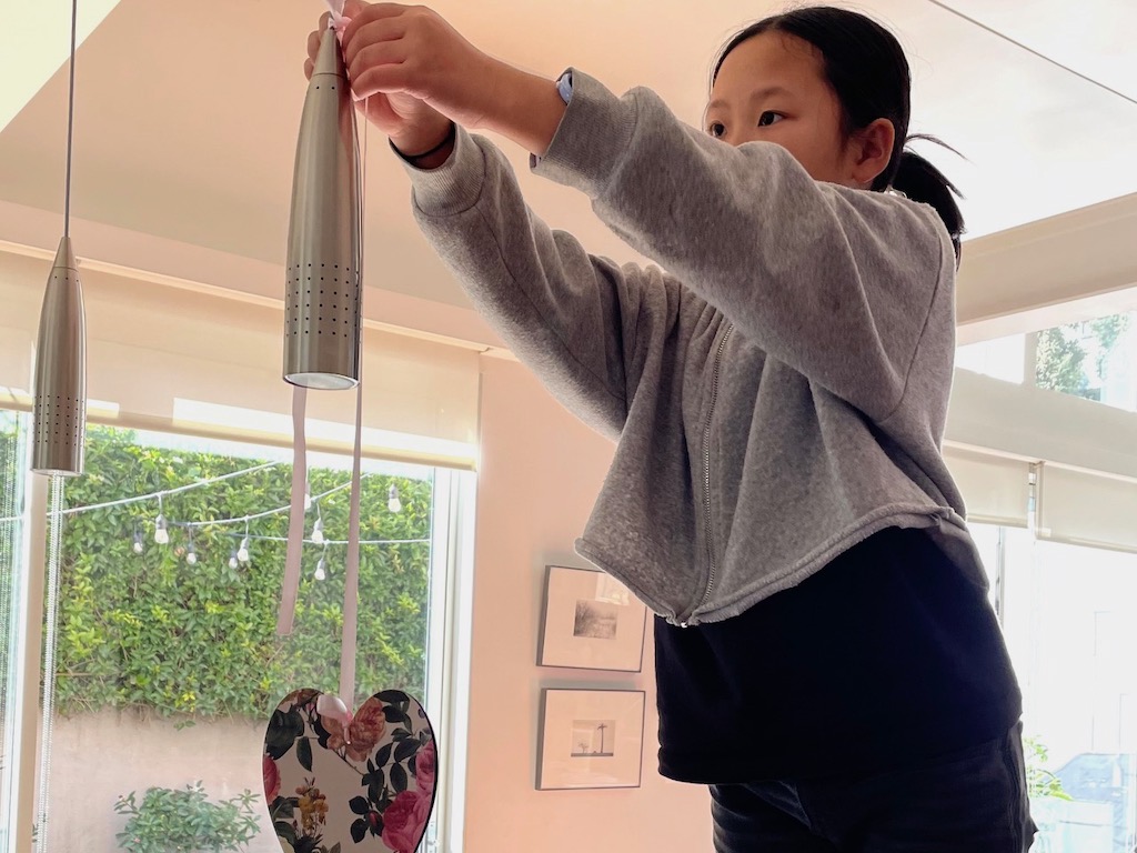 Child hangs a cardstock heart from a light fixture by tying it on with ribbons.