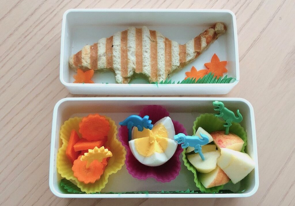 A brontosaurus bento box lunch is made by cutting a grilled cheese sandwich with a dinosaur cookie cutter.